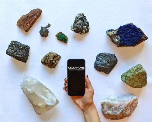 a hand holding a cellphone above a collection of rocks and mineral samples. The text on the cellphone screen reads "Cellphone: Unseen Connections"