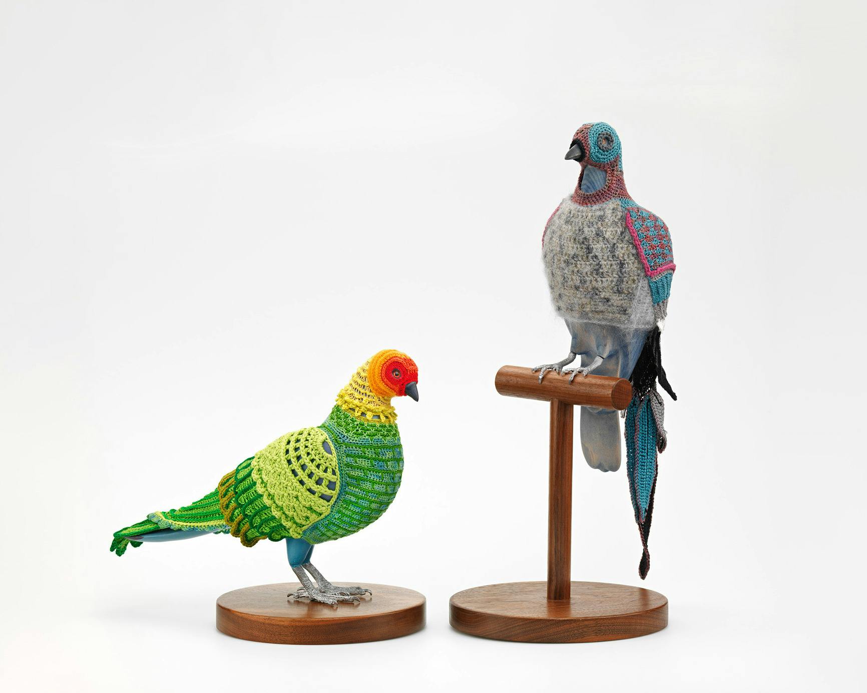 Two artworks, Biodiversity Reclamation Suits, by artist Laurel Roth Hope. Left: a crocheted suit with green, yellow, and red patterns on a pigeon mannequin. Right: another mannequin in a blue, gray, and red crochet suit. Both mannequins are mounted on round wooden bases."