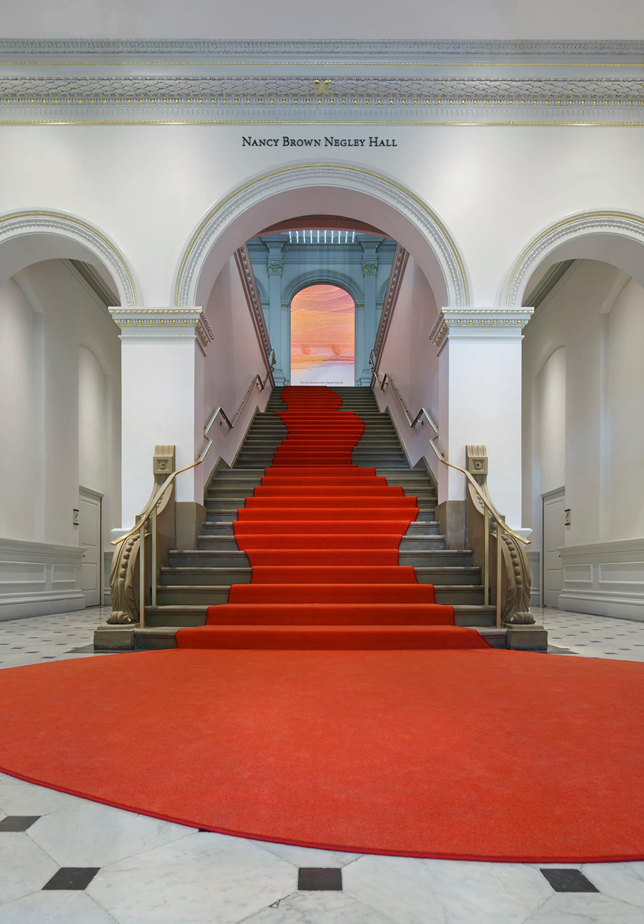 The grand staircase inside the Renwick Gallery, with a bright red carpet cascading down a grey stairway, flanked by three white archways
