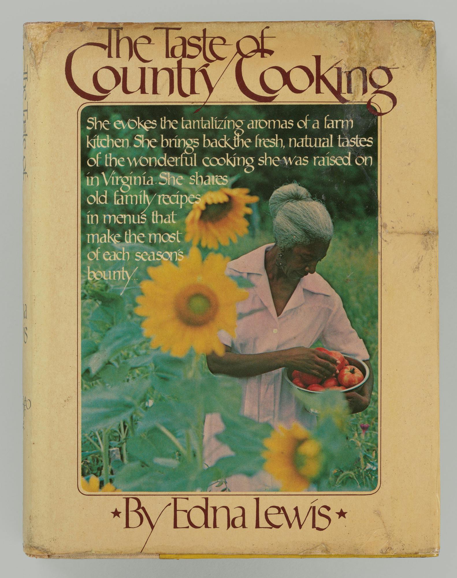 Cover of the book, The Taste of Country Cooking, by Edna Lewis. She is pictured holding a bowl of tomatoes in a field of sunflowers. Text overlays the photo: "She evokes the tantalizing aromas or a farm kitchen. She brings back the fresh, natural tastes of the wonderful cooking she was raised on in Virgnia. She shares old family recipes in menus that make the most of each season's bounty."