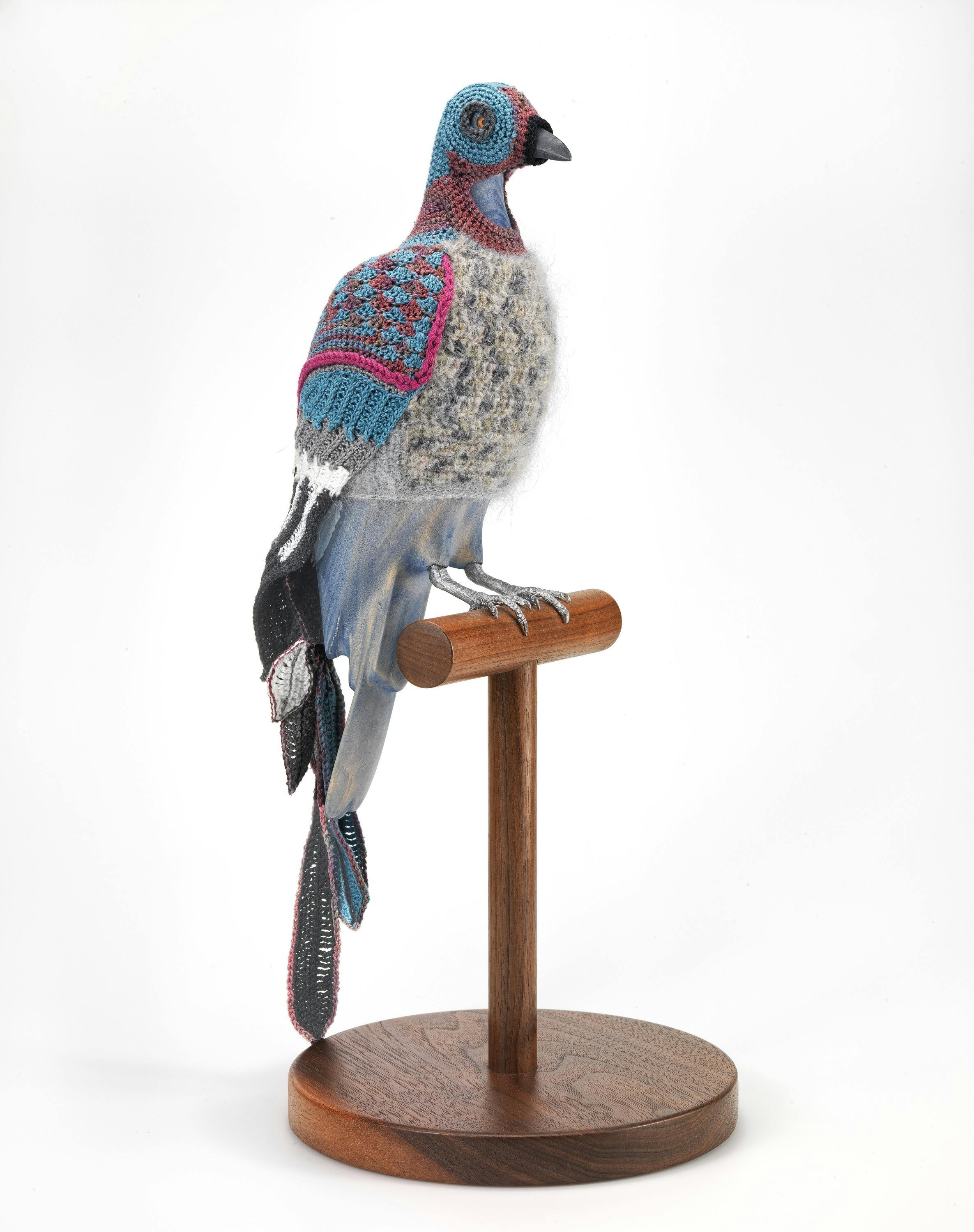 Model of a pigeon on a wooden pedestal wearing a bright blue, pink and grey knitted suit