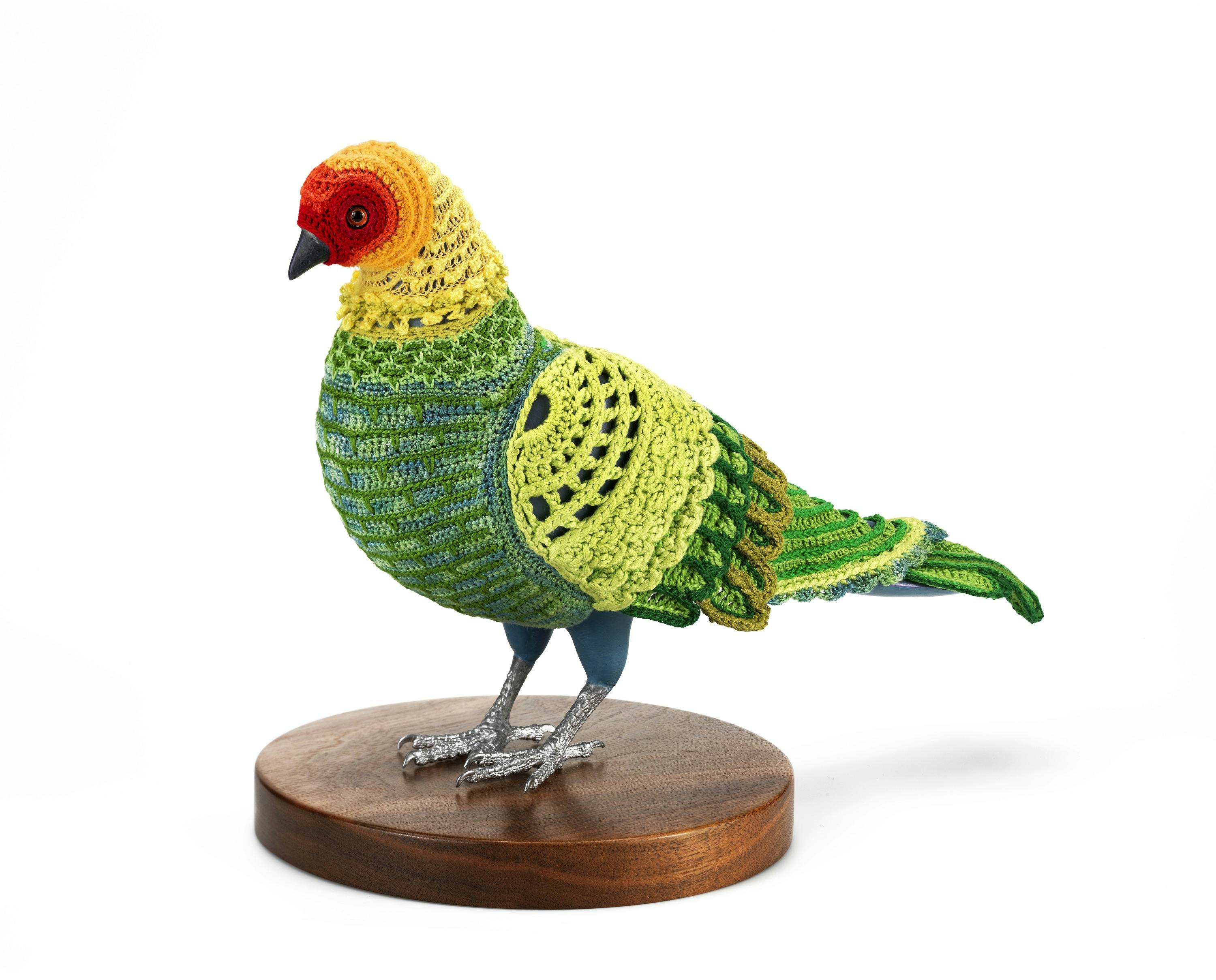 A crocheted suit with green, yellow, and red patterns on a pigeon mannequin, standing on a round wooden base